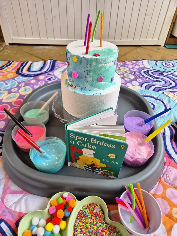 Cake decorating invitation themed on the picture book Spot Bakes a Cake: a plain two-tier play cake with straw candles, pots of coloured foam icing, pom poms and sprinkles.