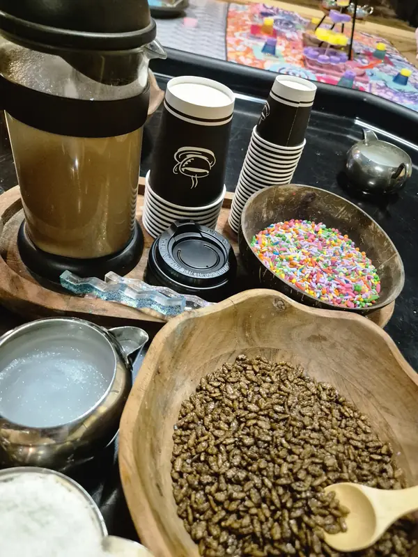 Pretend coffee shop set-up with takeaway cups and lids, a coffee plunger, a wooden bowl of brown sensory filler, sprinkles and sugar cubes.