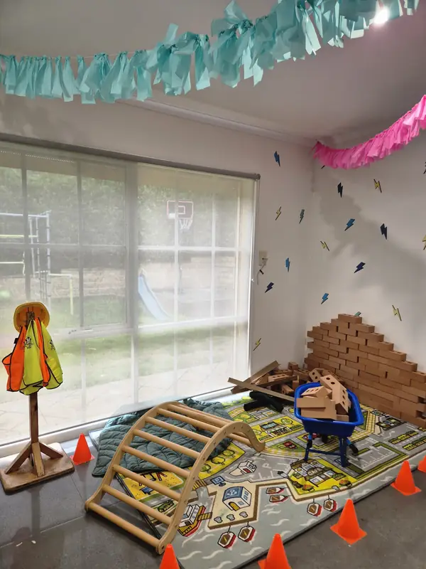 Indoor play corner set up for construction week with a climbing arch, hi-vis vests on a stand, cardboard bricks, traffic cones and a road play mat.