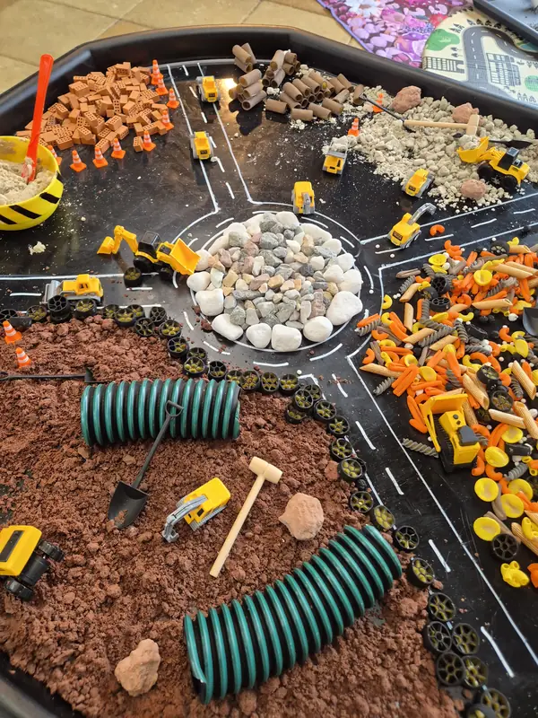 Construction-themed sensory tray with toy diggers, pebbles, pasta, tiny bricks and crumbly brown dirt laid out around a road circuit.