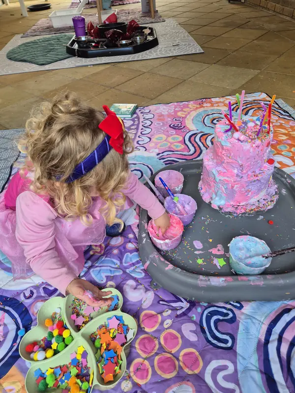 A little one in a pink tutu dips into pots of coloured foam while decorating a play cake, with a tray of pom poms and stars alongside.