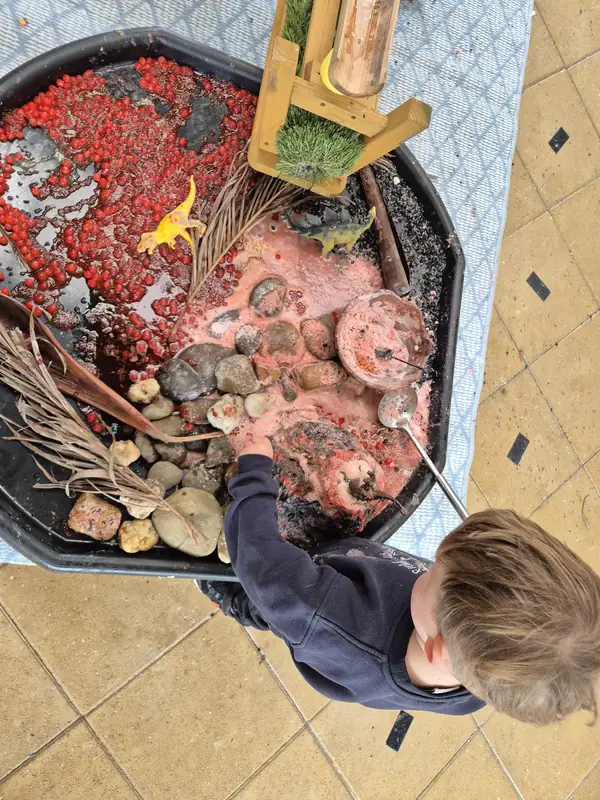 A child leans over a dinosaur tray full of red beads, stones and pink foam, stirring with a spoon.