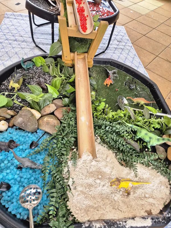 Dinosaur small-world tray with blue water beads, river stones, ferns, sand, toy dinosaurs and a bamboo chute.
