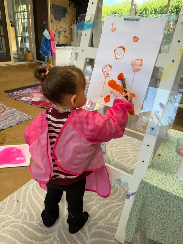 A little one in a pink smock paints orange circles on a big sheet of paper at an easel.