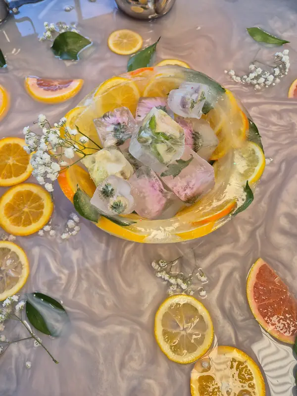 A bowl of ice blocks with flowers frozen inside, surrounded by citrus slices, leaves and sprigs of baby's breath floating in water.