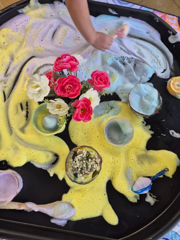 A child's hand reaches into a tray of yellow and white foam dotted with pink and white flowers and small bowls.