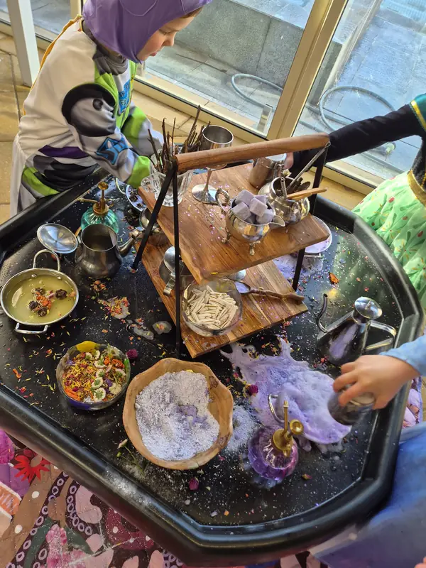 Children mix potions together at the potion-making tray, pouring from silver teapots into bowls of petals and powder.