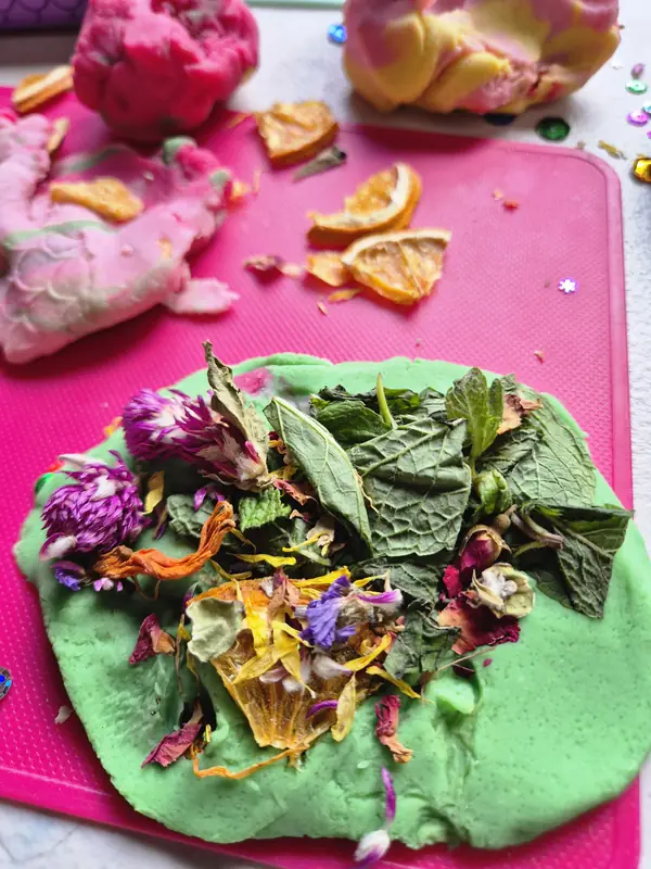 Green playdough pressed with mint leaves, dried flowers and petals, with pink and yellow playdough and dried orange slices nearby.