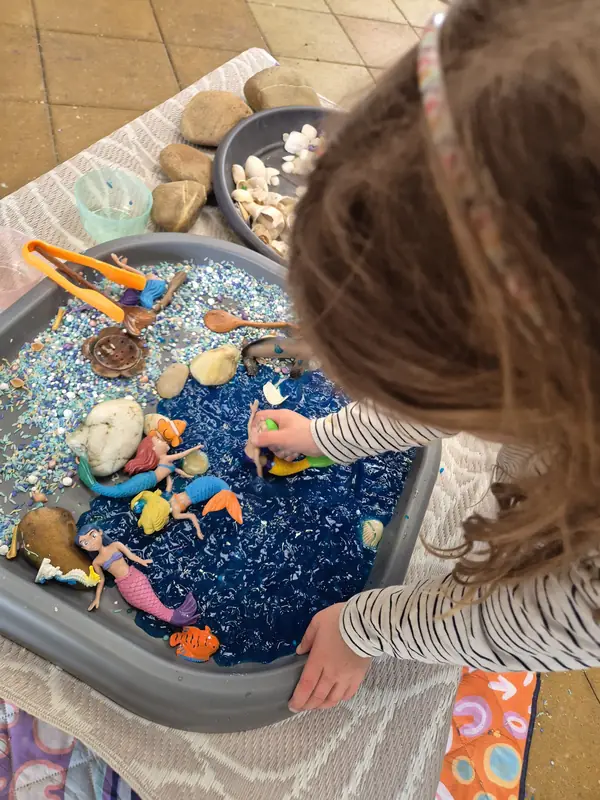 A child scoops blue water beads in an ocean sensory tray with shells, sand and mermaid figures.