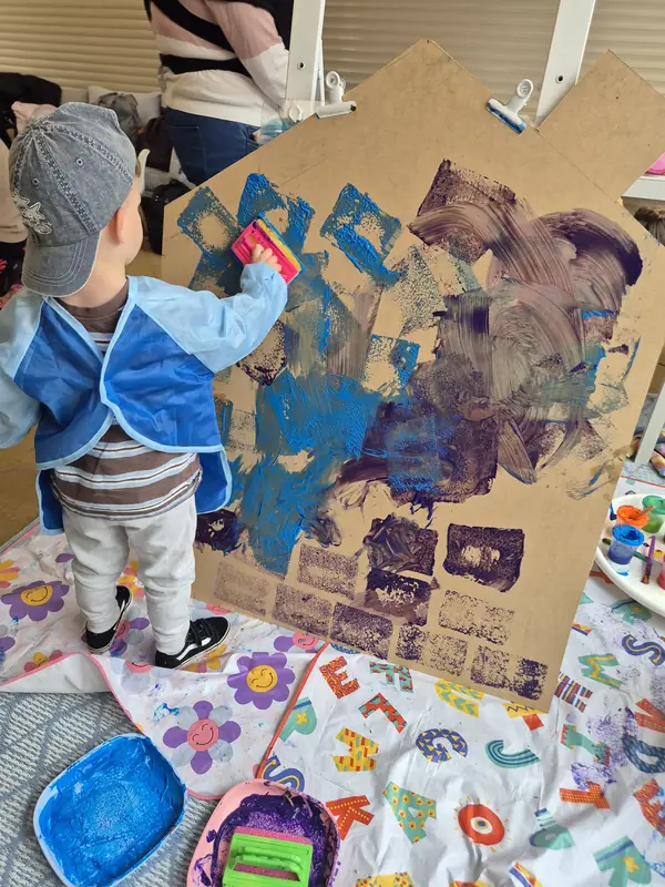 A little one in a blue smock and cap stamps blue and purple paint onto a big cardboard house.