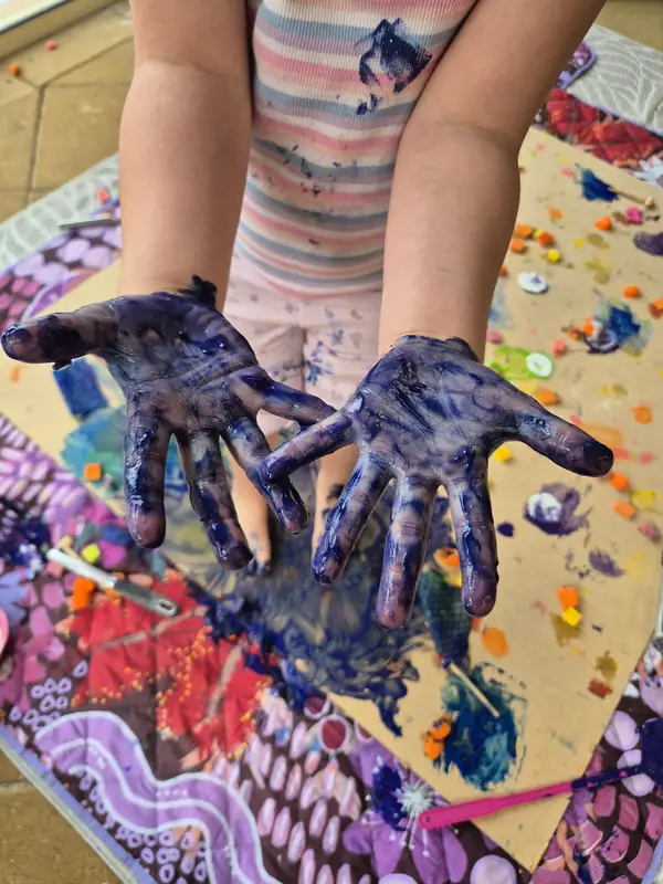 A child holds out two hands covered in blue and purple paint above a messy painting mat.