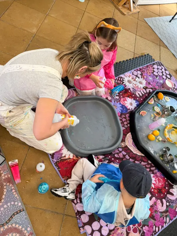 A grown-up kneels on the play mat helping two little ones squeeze colour into a tray, with a citrus sensory tray beside them.