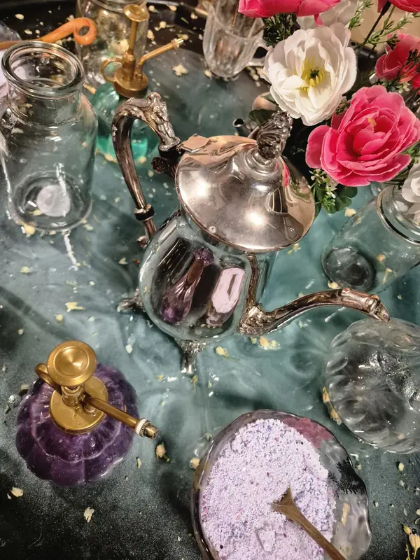Close-up of a potions table with a silver teapot, glass jars, a purple perfume bottle, lavender-coloured powder and silk flowers.