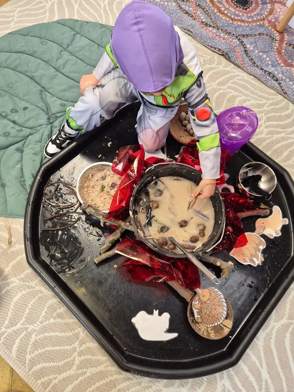 A child in a space ranger costume kneels on the Wombat Stew tray and stirs a muddy stew in the big pot.