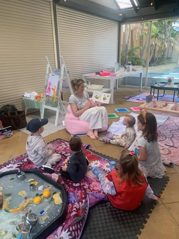 Story time in the undercover outdoor area: a grown-up reads a picture book to a small group of children sitting on the play mats.