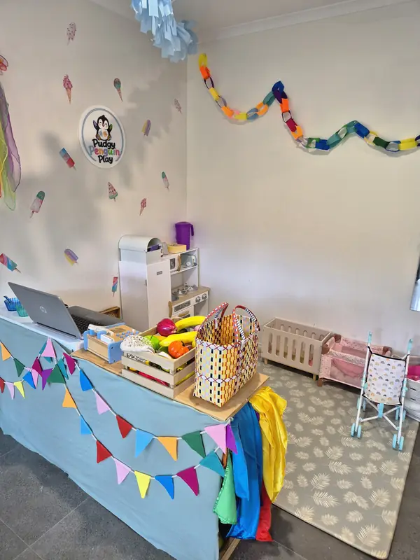The home studio set up with a play shop counter, bunting, baskets of pretend fruit, a toy kitchen and a dolls' corner, with the Pudgy Penguin Play logo on the wall.
