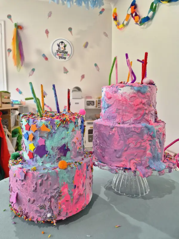 Two decorated two-tier play cakes covered in pink, purple and blue foam, sprinkles and pipe-cleaner candles, on display in the studio.