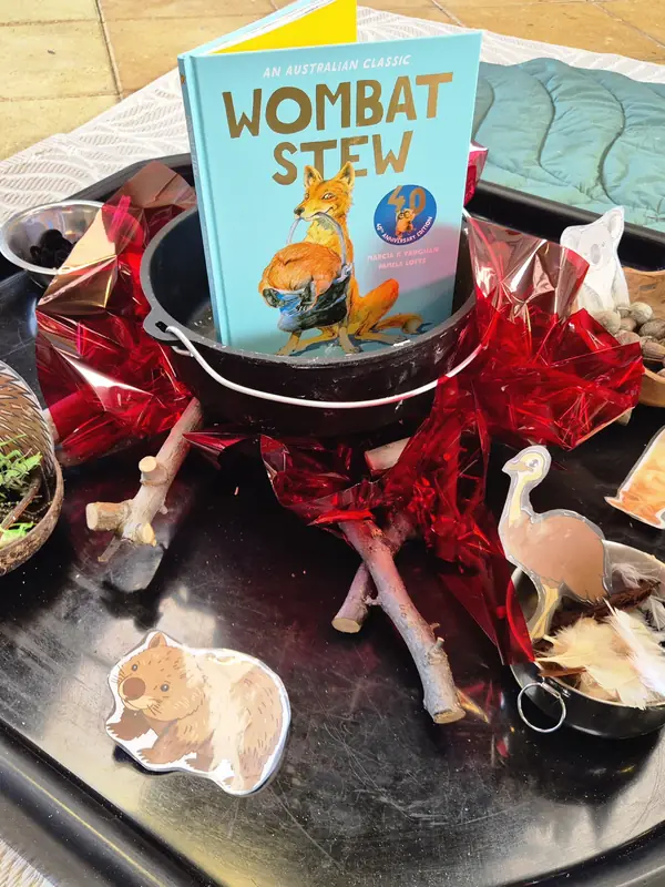 Story tray themed on the picture book Wombat Stew, with the book standing in a cooking pot surrounded by sticks, gumnuts, feathers and Australian animal cut-outs.
